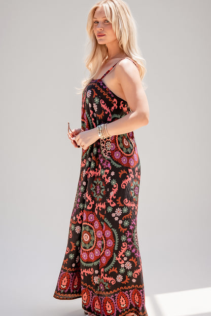 A woman modeling a sleeveless maxi dress with a black base and a vibrant, multicolored bohemian paisley and floral pattern, set against a plain light gray studio background.