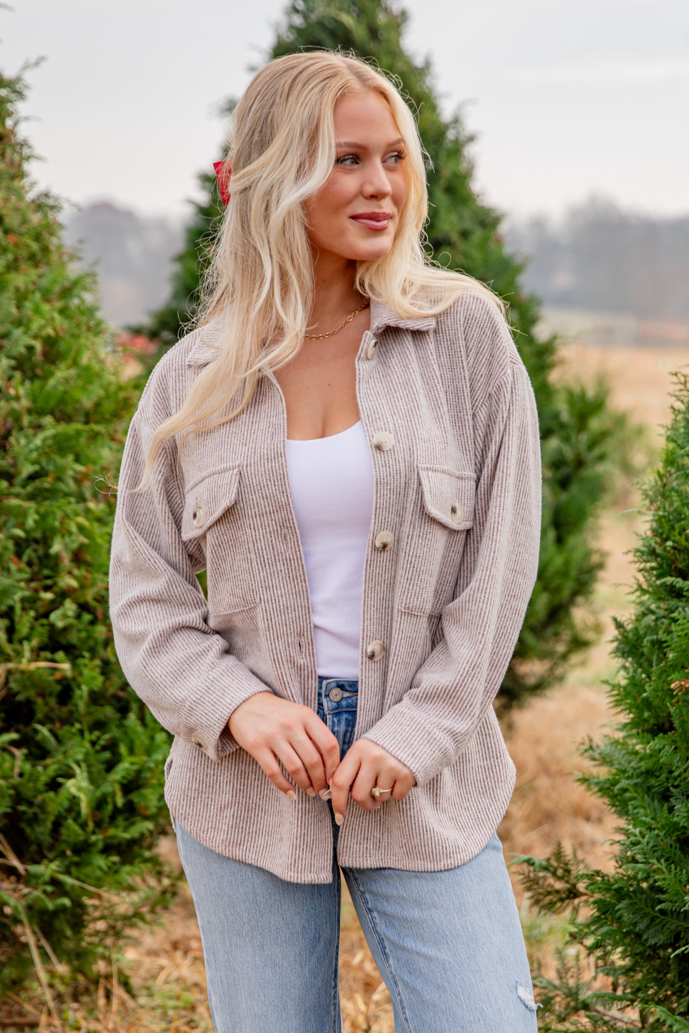 Woman standing in a Christmas tree farm wearing a beige jacket and light blue jeans.
