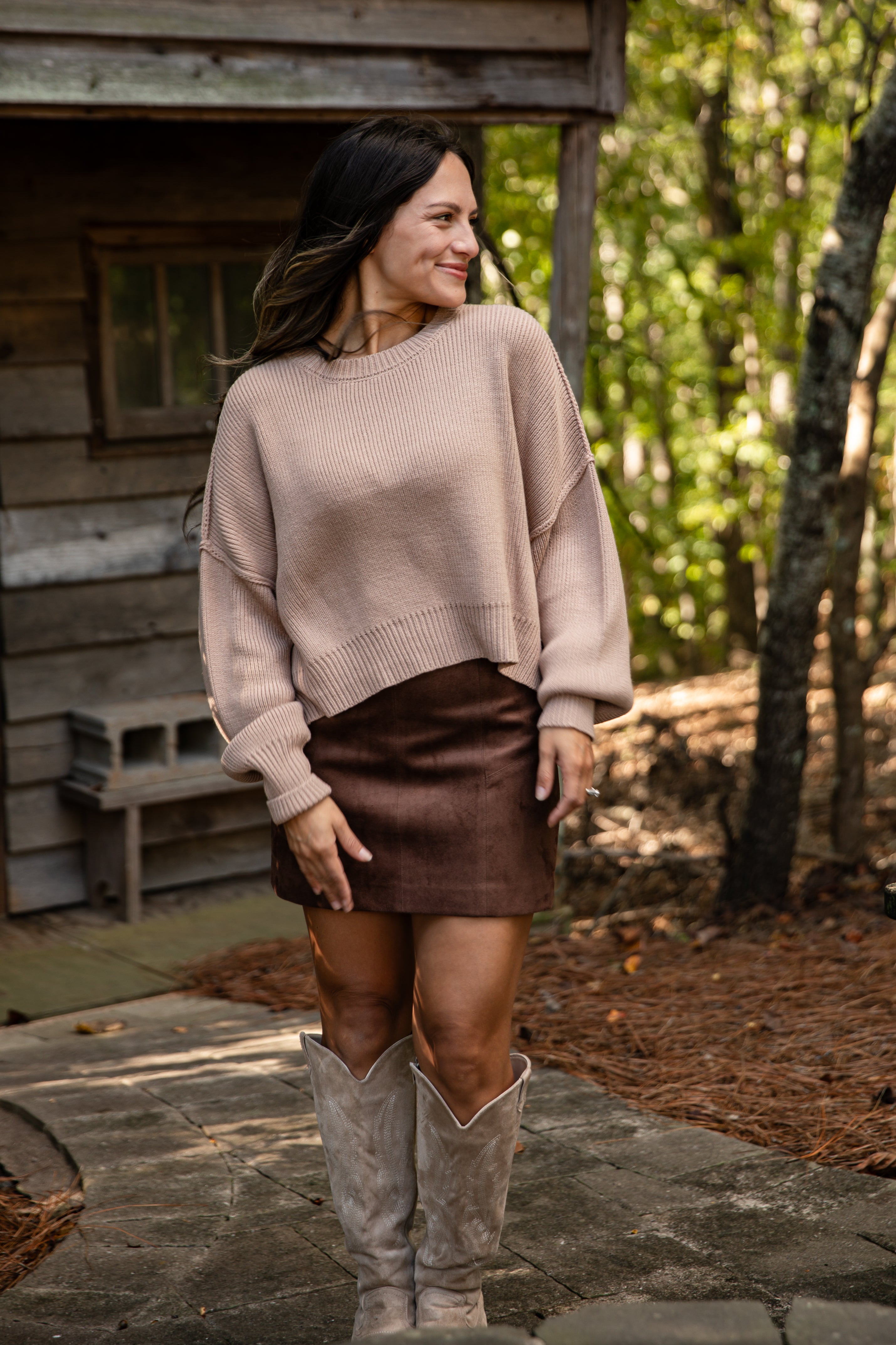 Woman in a beige sweater and brown skirt standing in front of a wooden cabin.