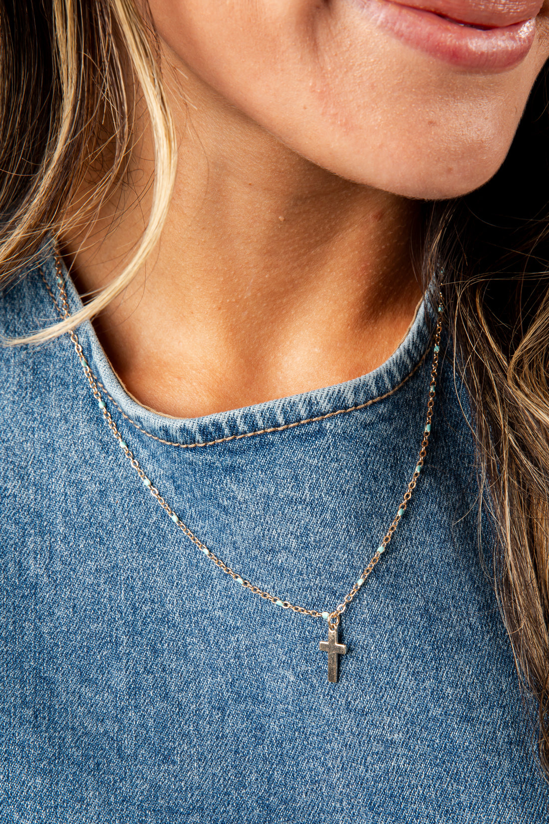 Close-up of a person wearing a silver necklace with a cross pendant on a blue denim background.