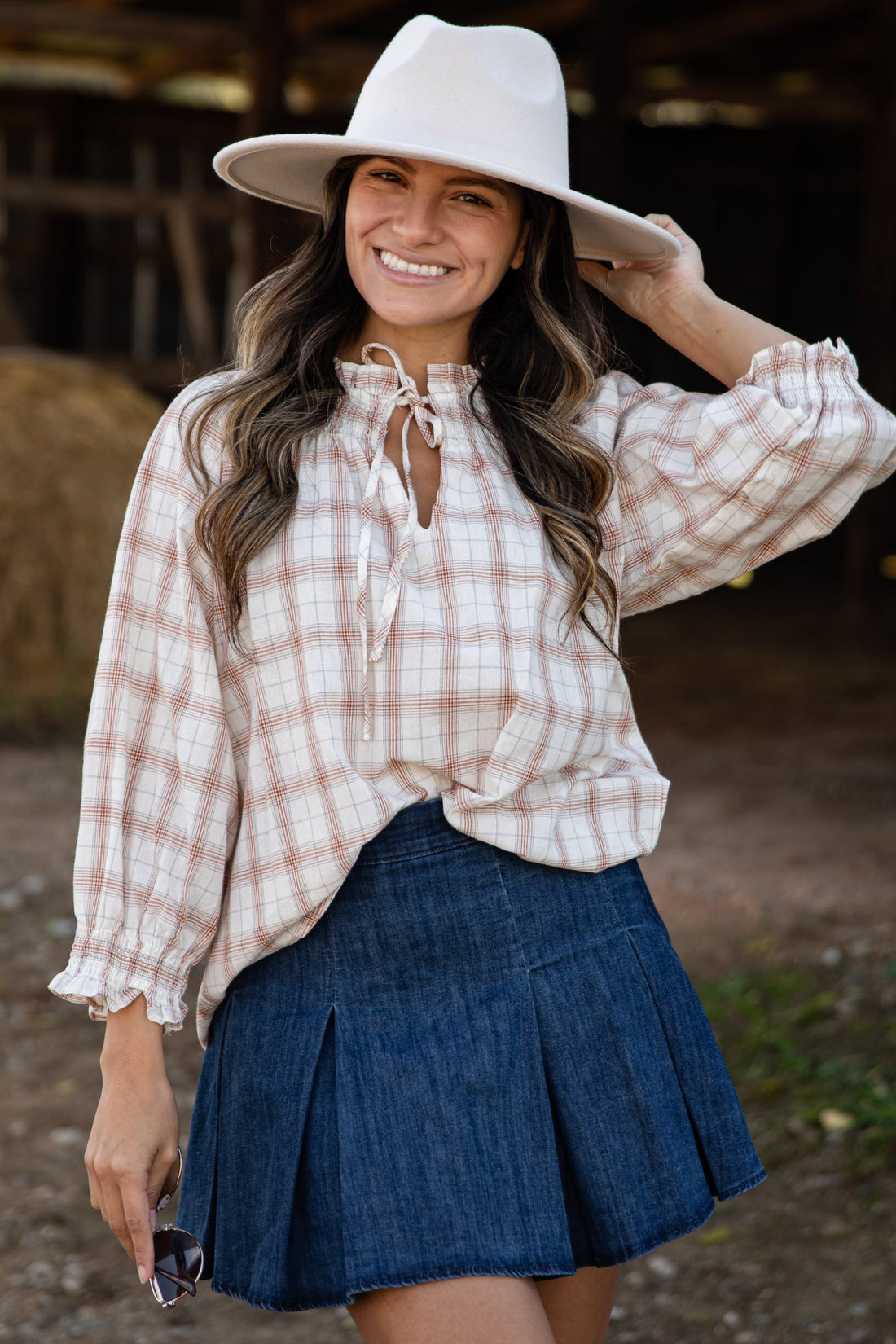 Woman wearing a plaid shirt, denim skirt, and white hat in a rustic setting.