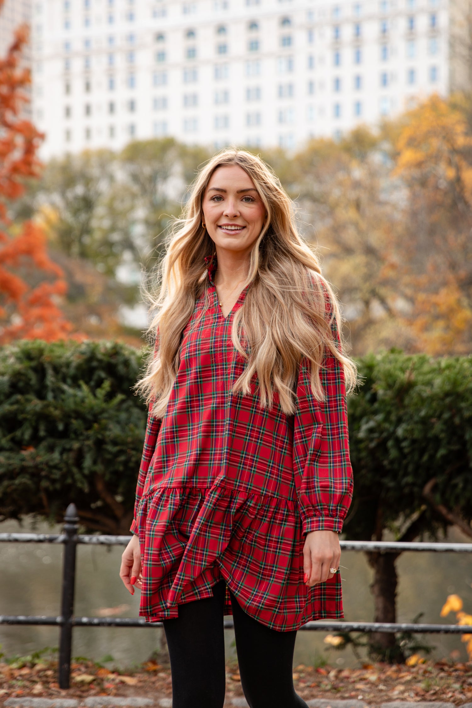 Woman wearing a red plaid dress standing in an urban park with trees in the background