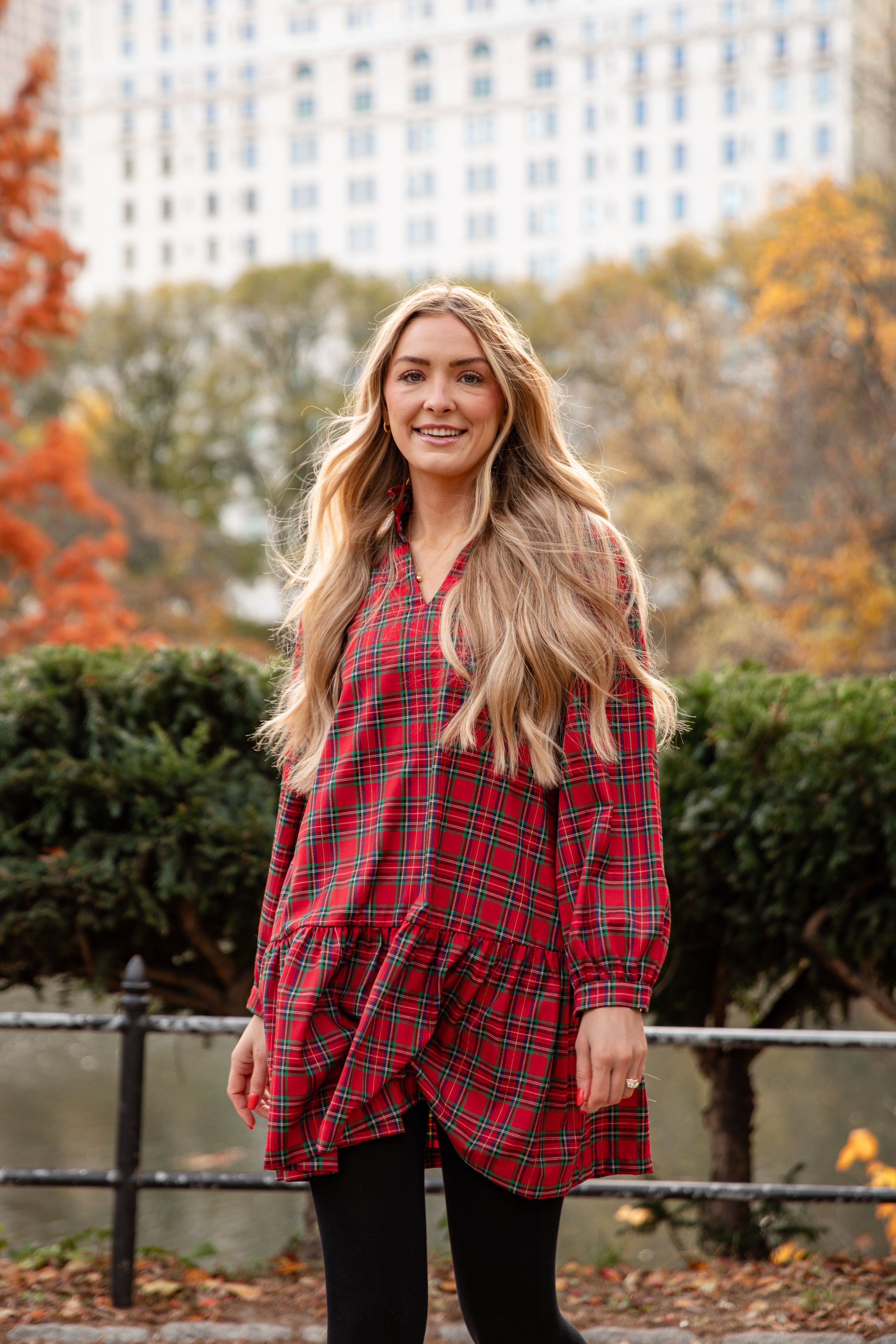 Woman wearing a red plaid dress standing in an urban park with trees in the background