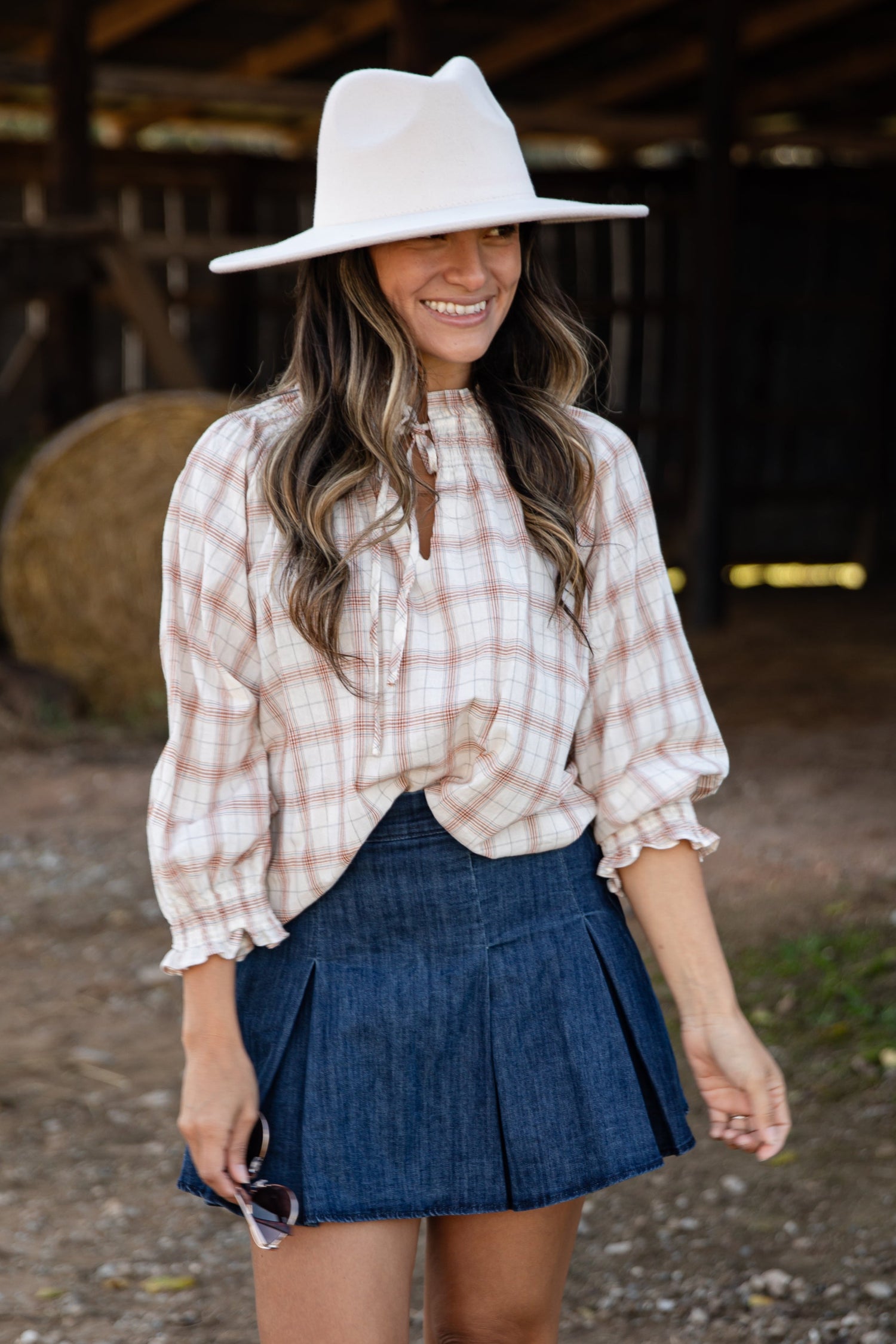 Woman wearing a plaid shirt, denim skirt, and cowboy boots in a barn setting.