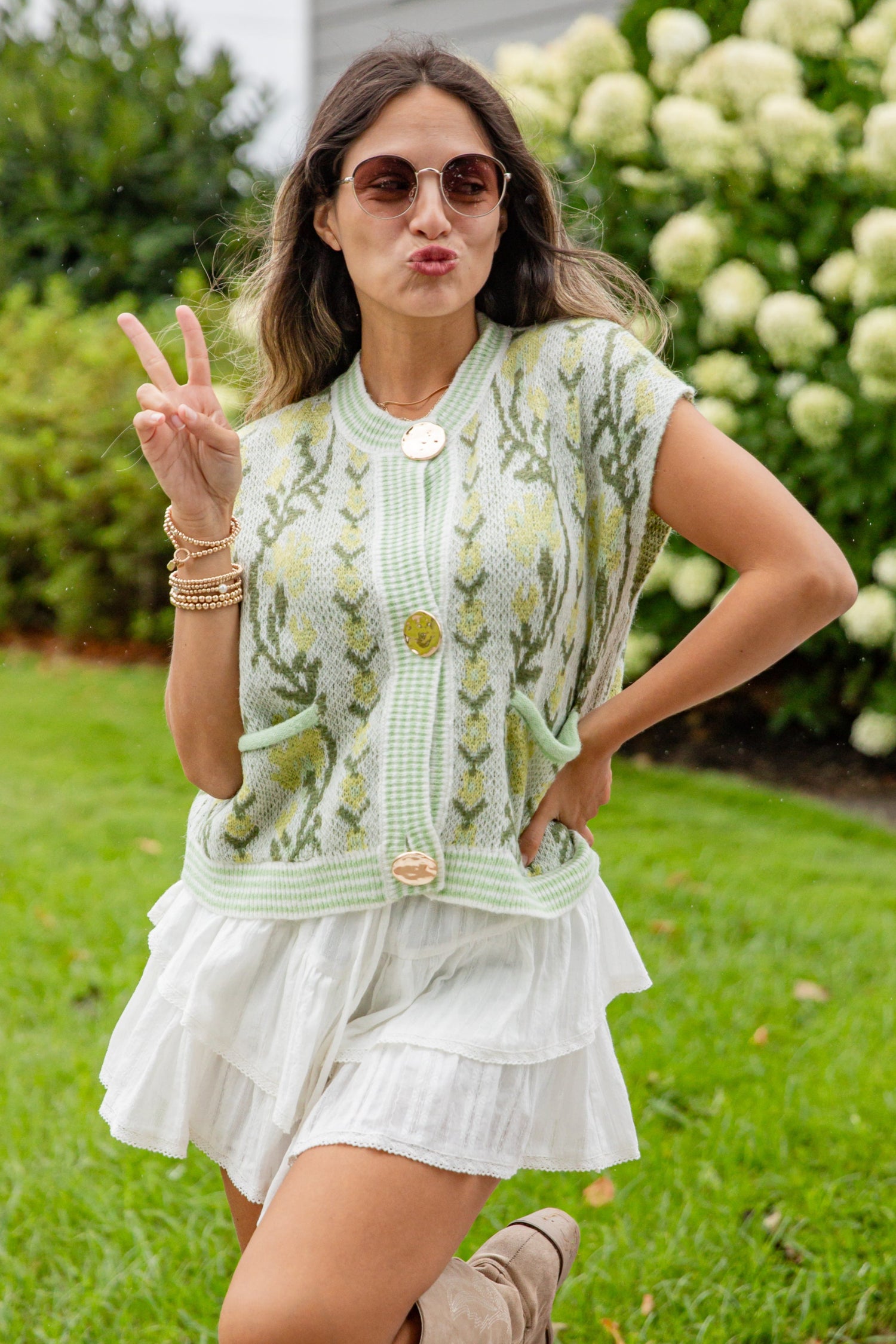 Woman in a green floral cardigan and white dress standing outdoors with flowers in the background