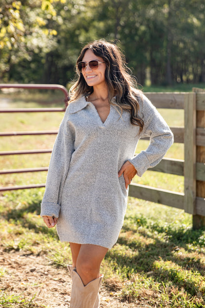 Woman wearing a gray sweater dress and beige boots standing in a field with a wooden fence and trees in the background.
