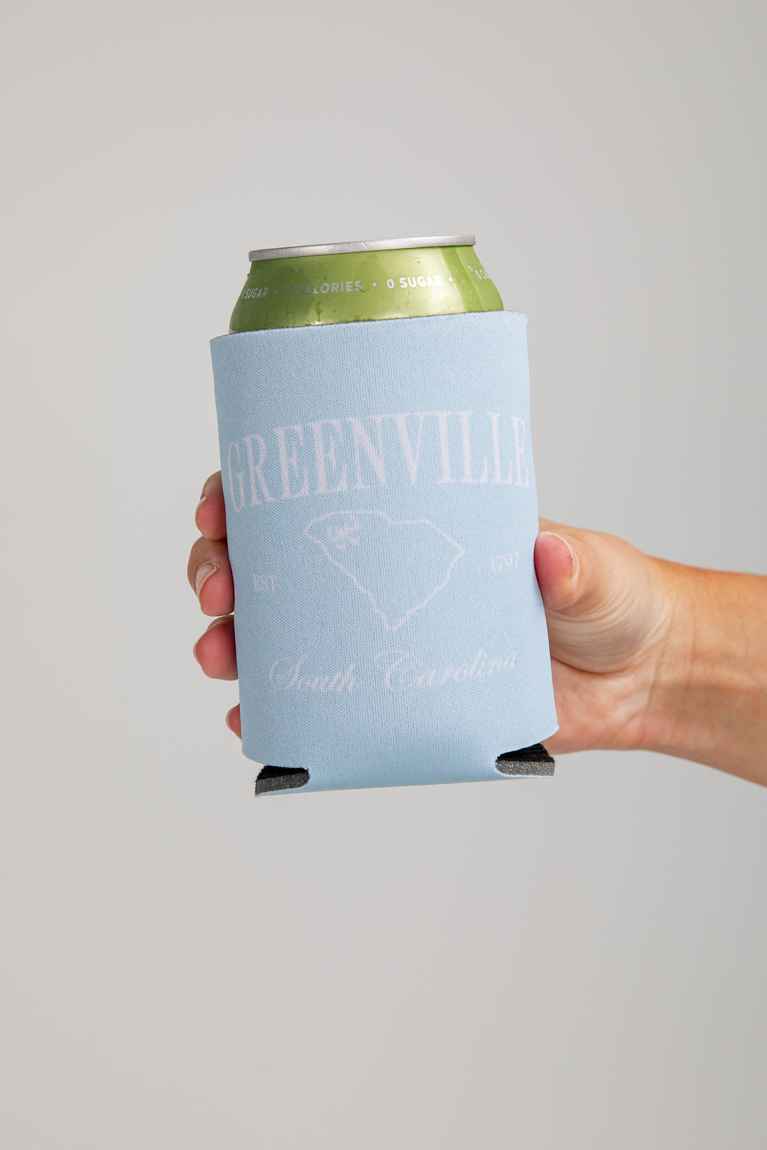 Hand holding a can with a Greenville, South Carolina branded koozie against a gray background
