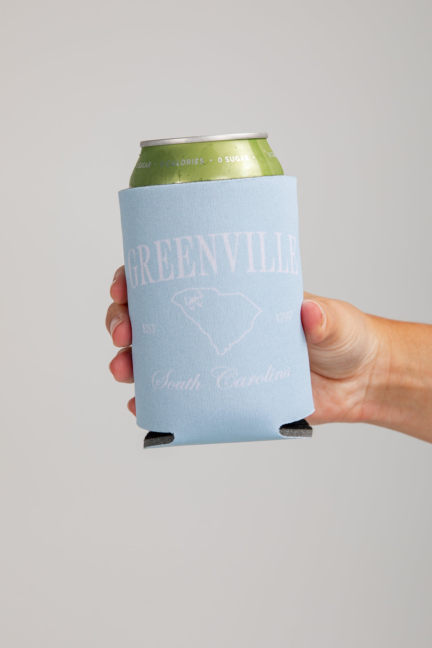 Hand holding a can with a Greenville, South Carolina branded koozie against a gray background