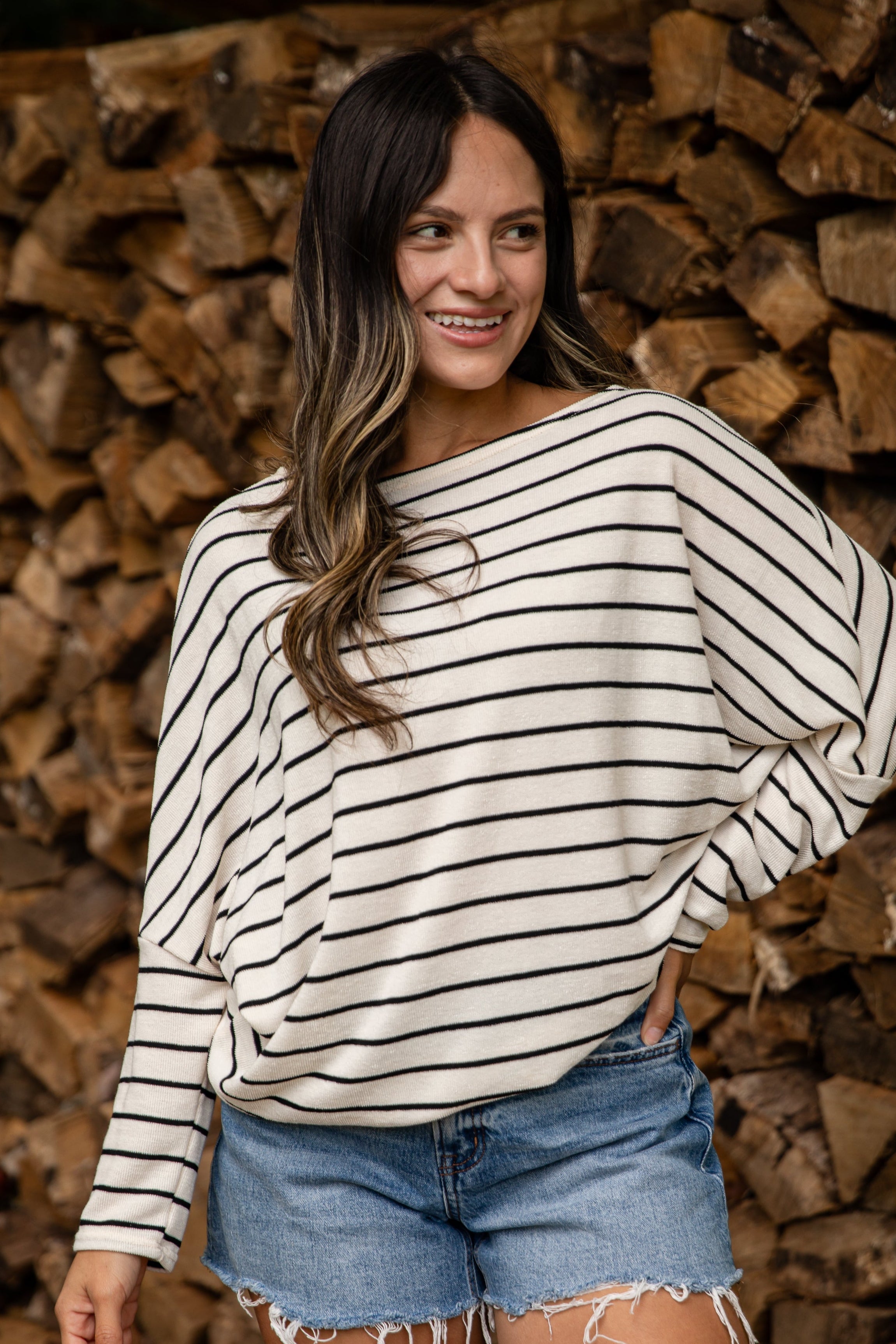 Woman wearing a striped sweater and denim shorts standing in front of stacked wood.
