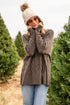 Woman in a gray coat and white beanie standing in a Christmas tree lot.
