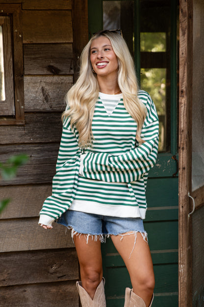 Woman wearing a green and white striped sweater and denim shorts standing in front of a wooden cabin.