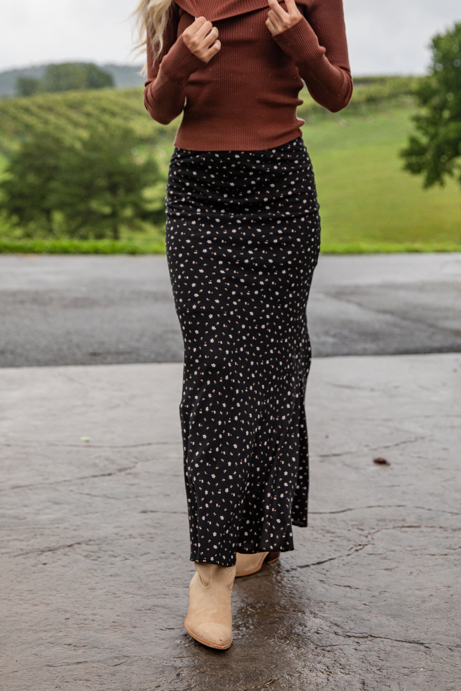 Person wearing a brown sweater and black floral skirt standing on a paved surface with greenery in the background.