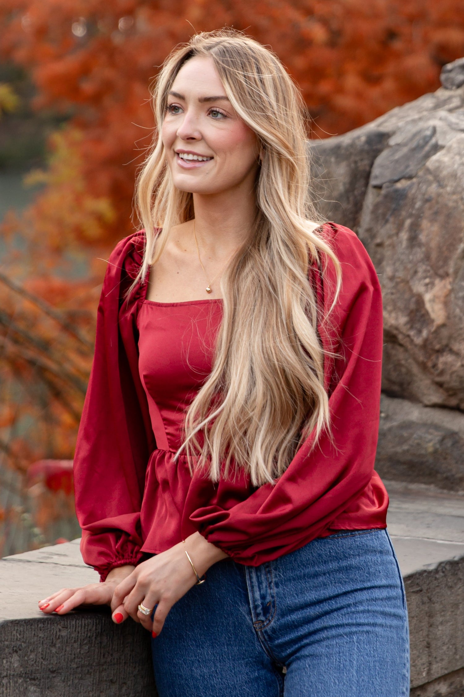 Woman in a red top and blue jeans leaning against a stone wall with autumn trees in the background