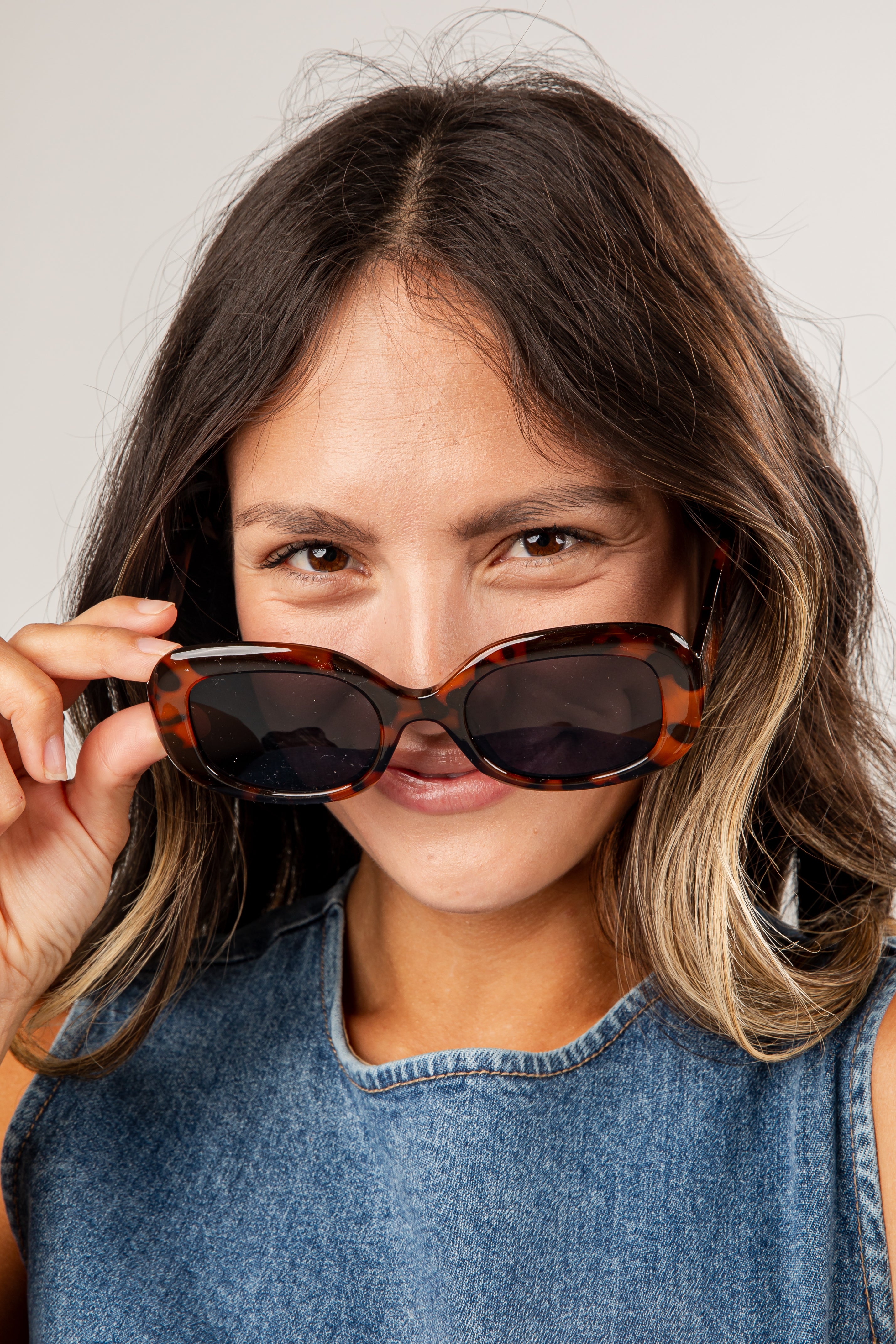 Woman holding tortoiseshell sunglasses in front of her face against a plain background
