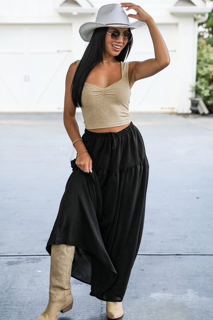 Woman wearing a beige top, black wide-leg pants, and a white hat in an outdoor setting.