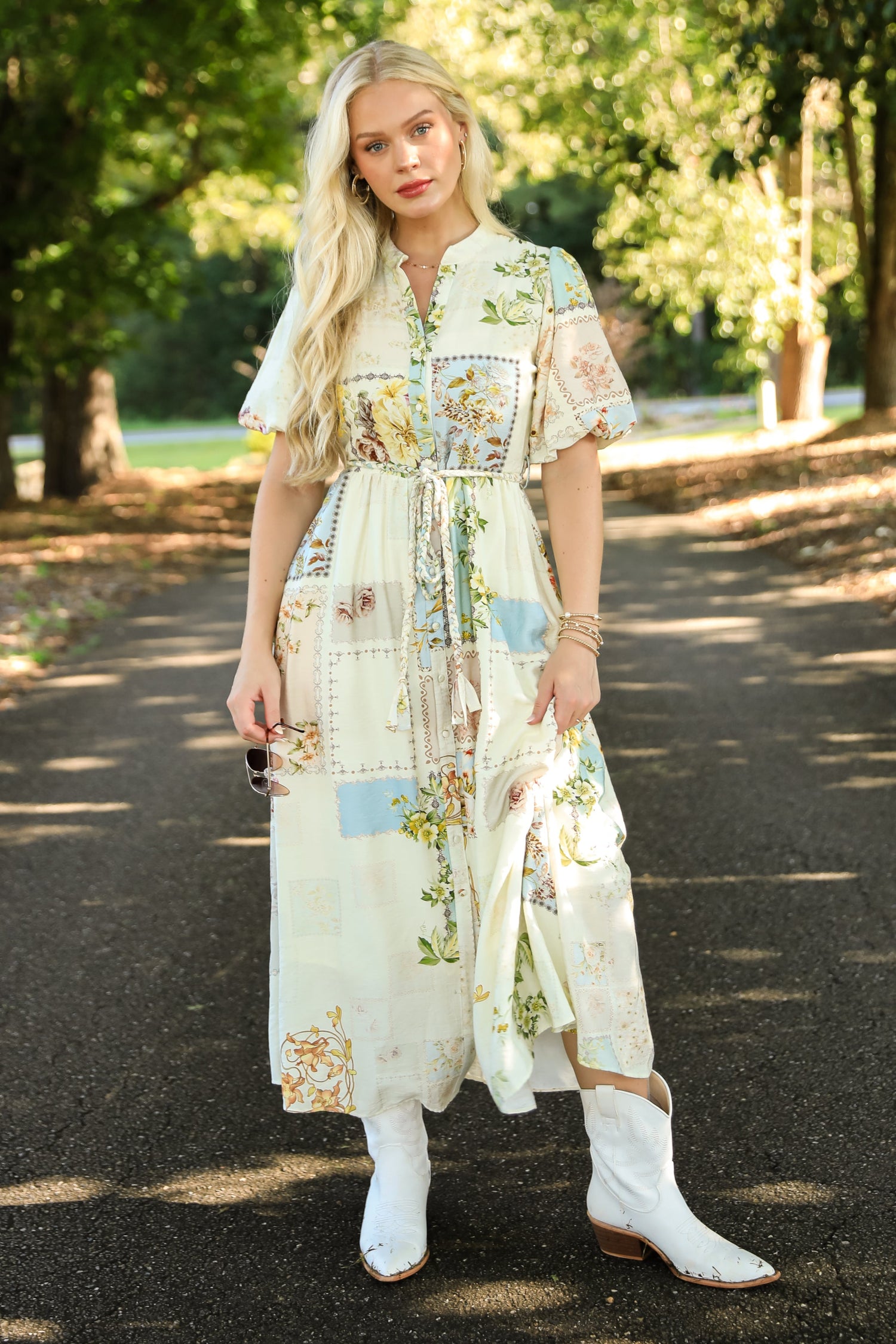 Woman in a floral dress and white boots standing on a tree-lined path.