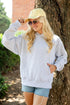 A woman wearing a light gray oversized hoodie, denim shorts, a yellow baseball cap, and sunglasses stands outdoors next to a large tree trunk.
