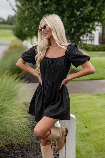 Woman in a black dress and beige boots standing outdoors with greenery in the background
