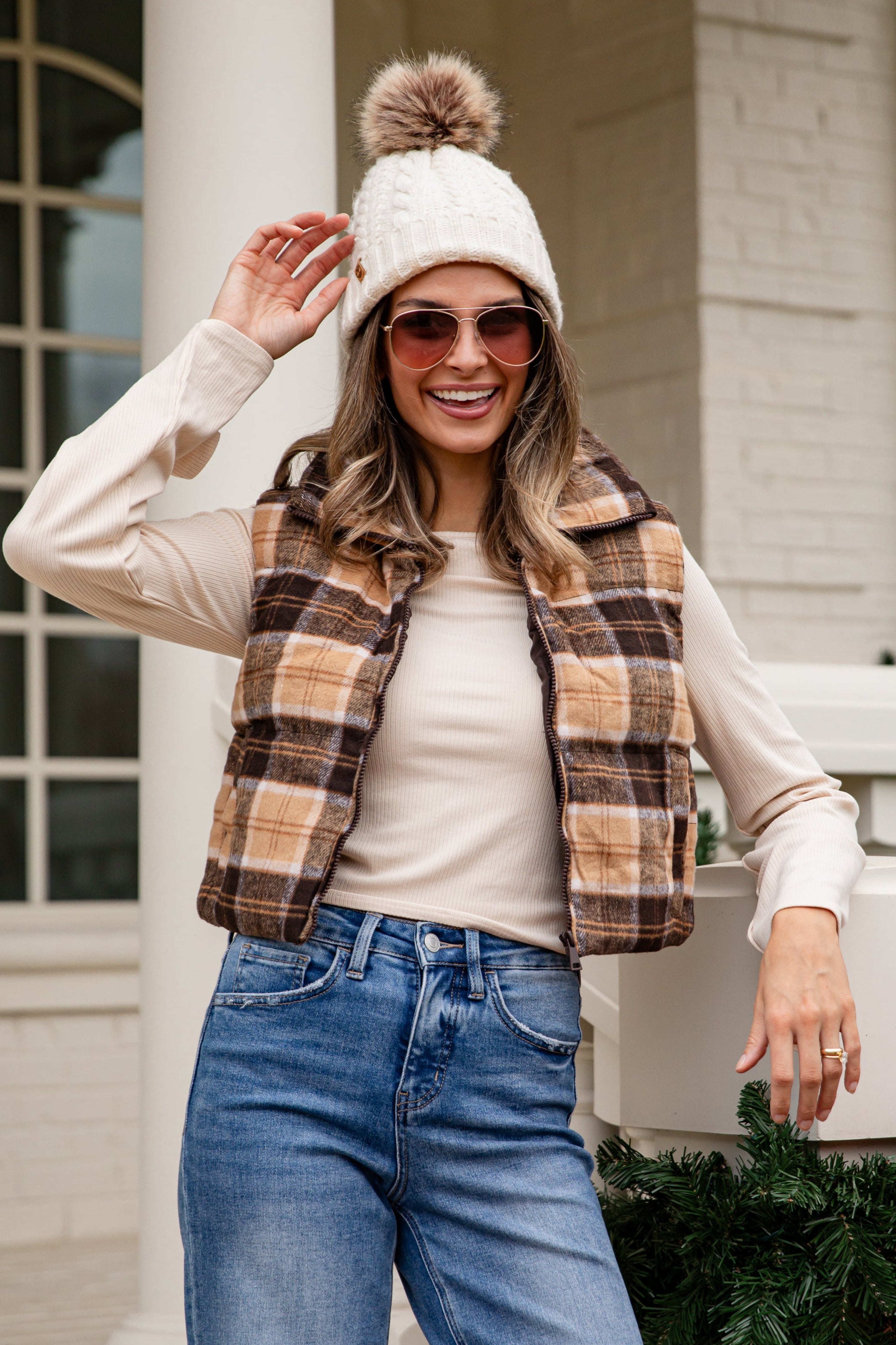 Woman wearing a plaid jacket, white beanie with pom-pom, and sunglasses, standing on a porch.