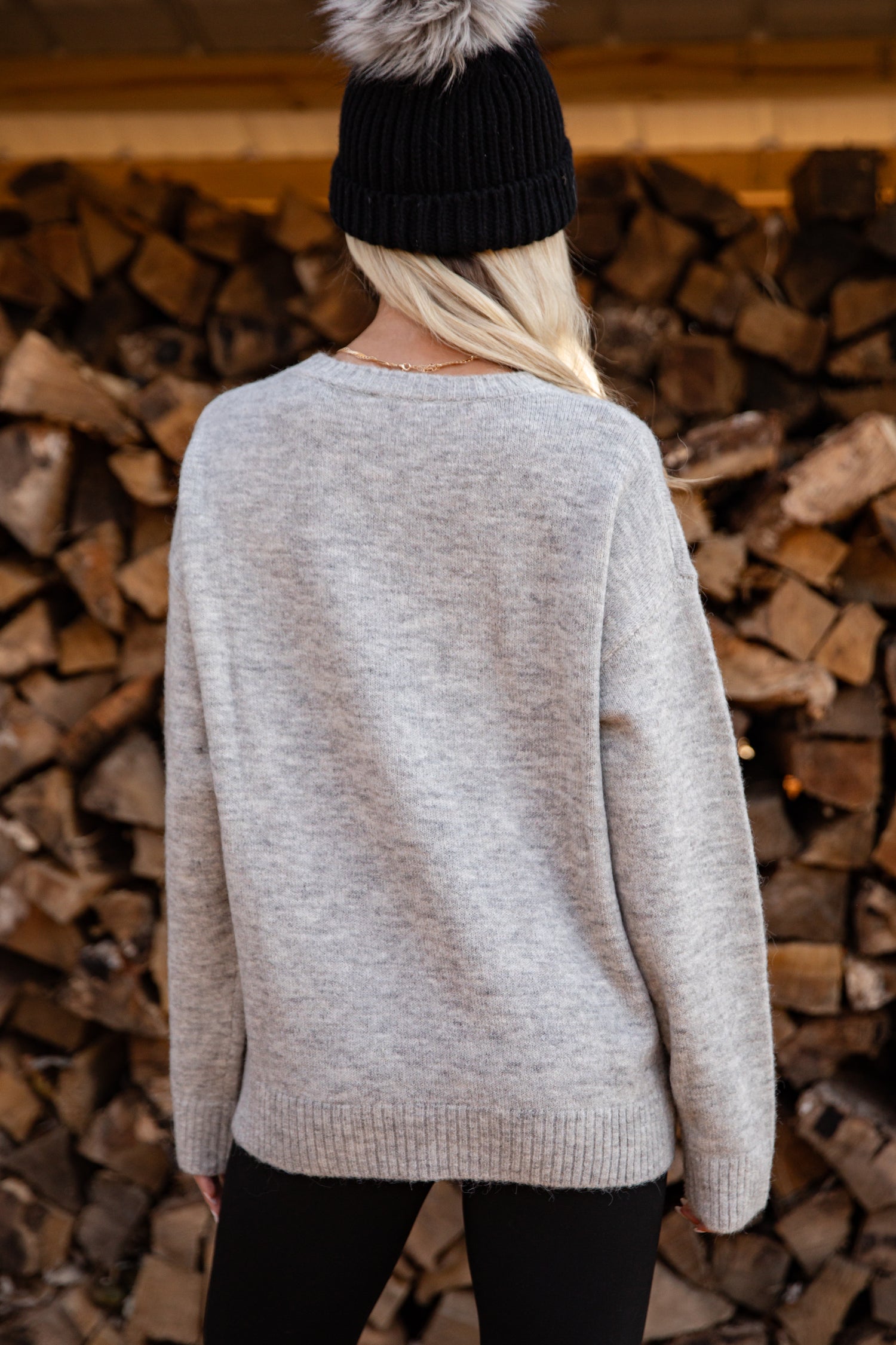Person wearing a light gray sweater and black beanie standing in front of stacked firewood.