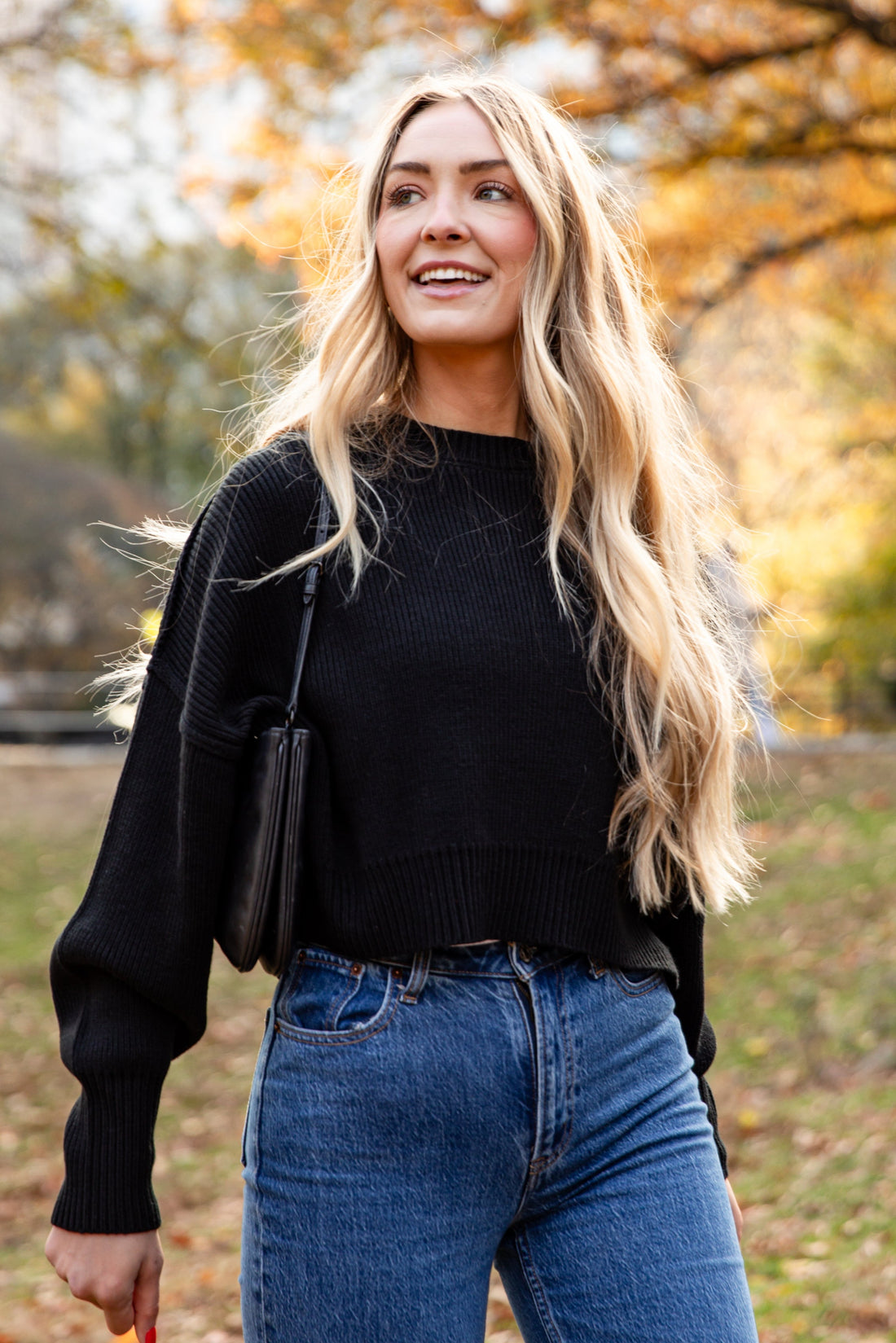 Woman in black sweater and blue jeans standing in a park with autumn trees.