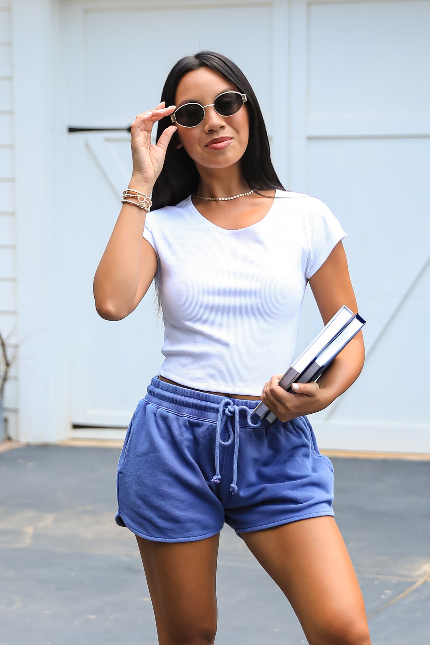 Woman wearing a white t-shirt and blue shorts, holding sunglasses and a book.