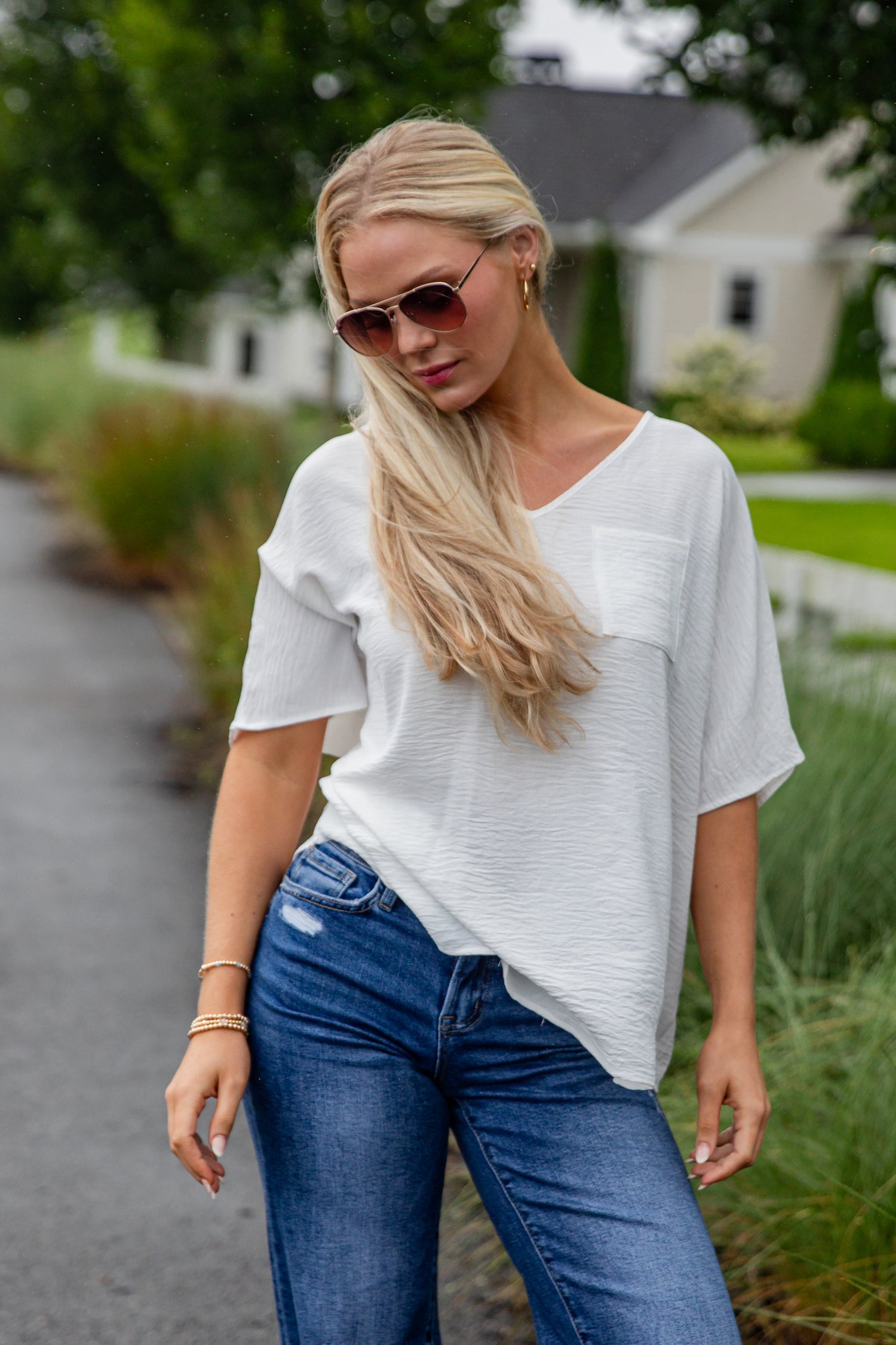 Woman wearing a white top and blue jeans standing outdoors with trees and a house in the background.