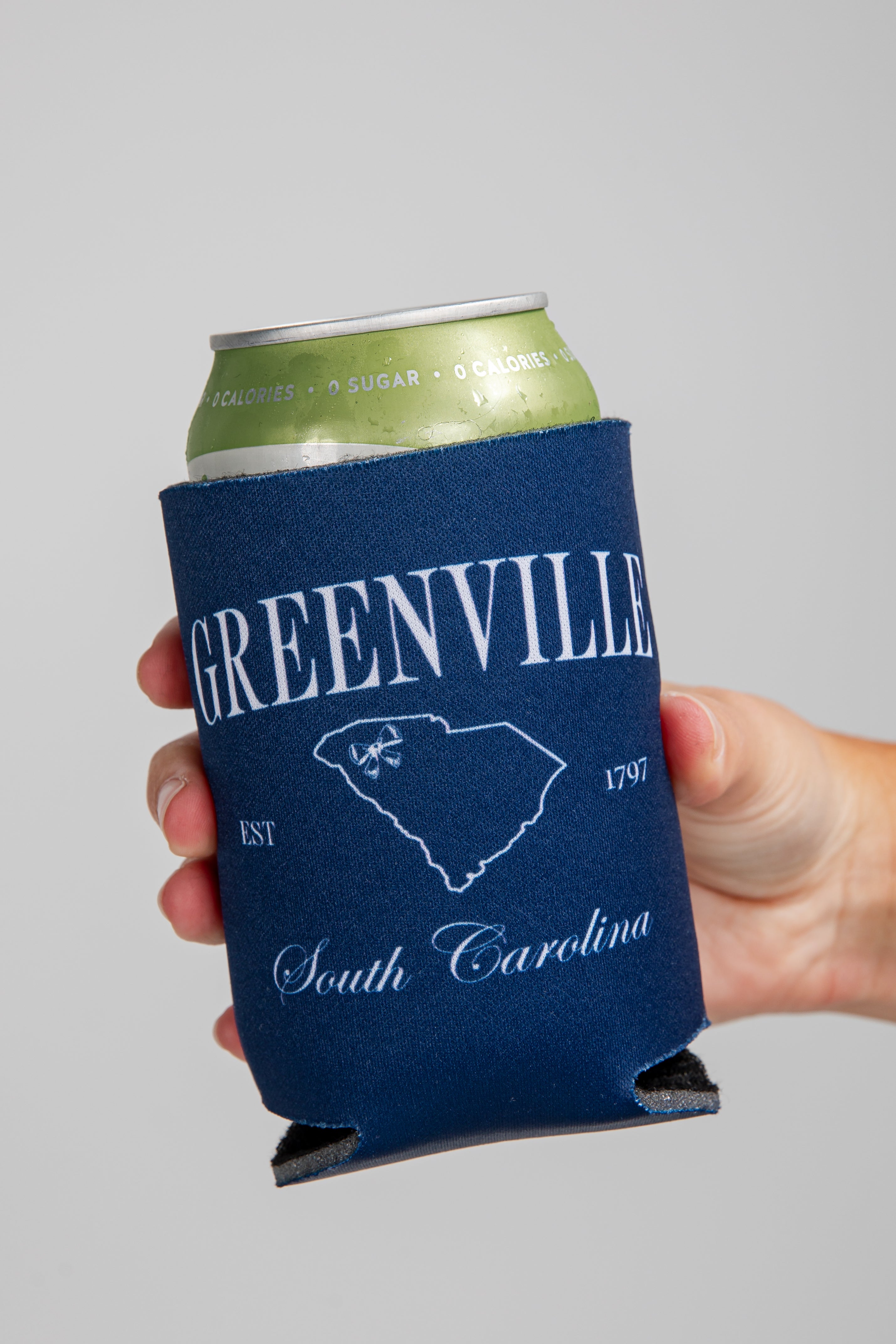 Navy blue can cooler with Greenville, South Carolina design held by a hand against a gray background