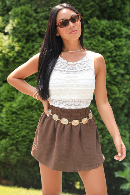 Woman wearing a white sleeveless top and brown skirt with sunglasses against a green hedge background