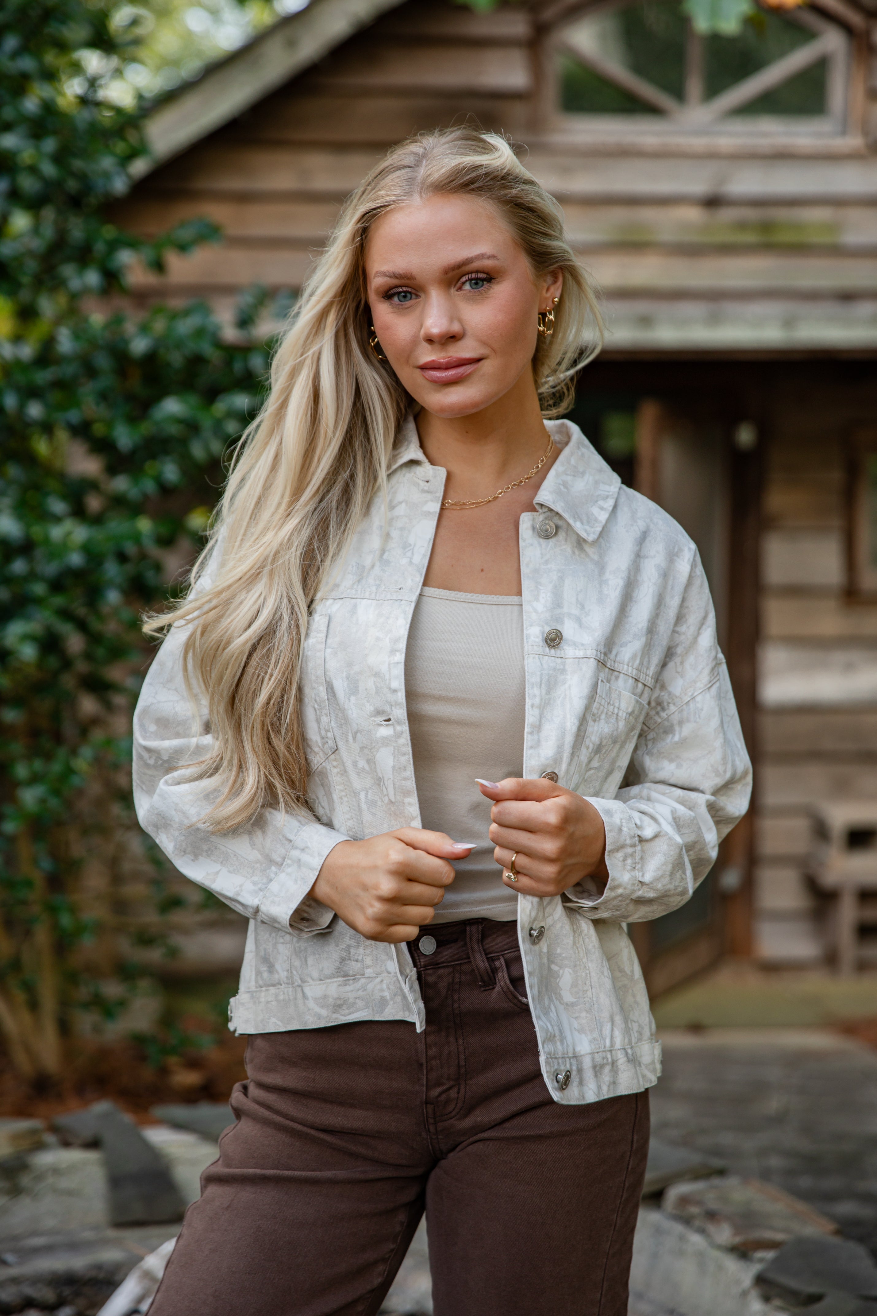 Woman wearing a light-colored jacket and brown pants standing outdoors with a wooden structure and greenery in the background.