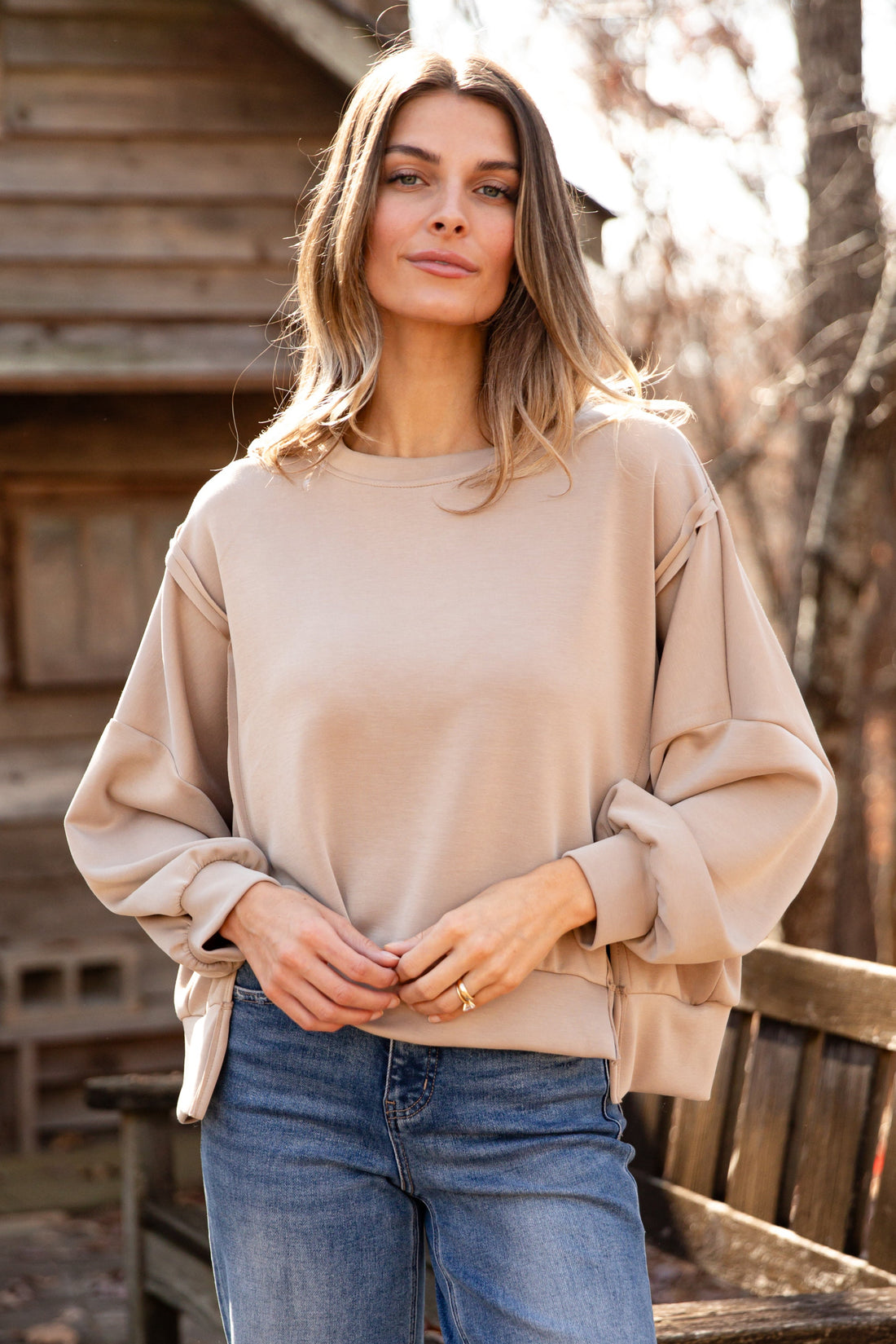 Woman wearing a beige sweater and blue jeans standing outdoors near a wooden cabin.