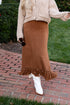 Person wearing a beige cardigan and brown fringed skirt on a brick path.