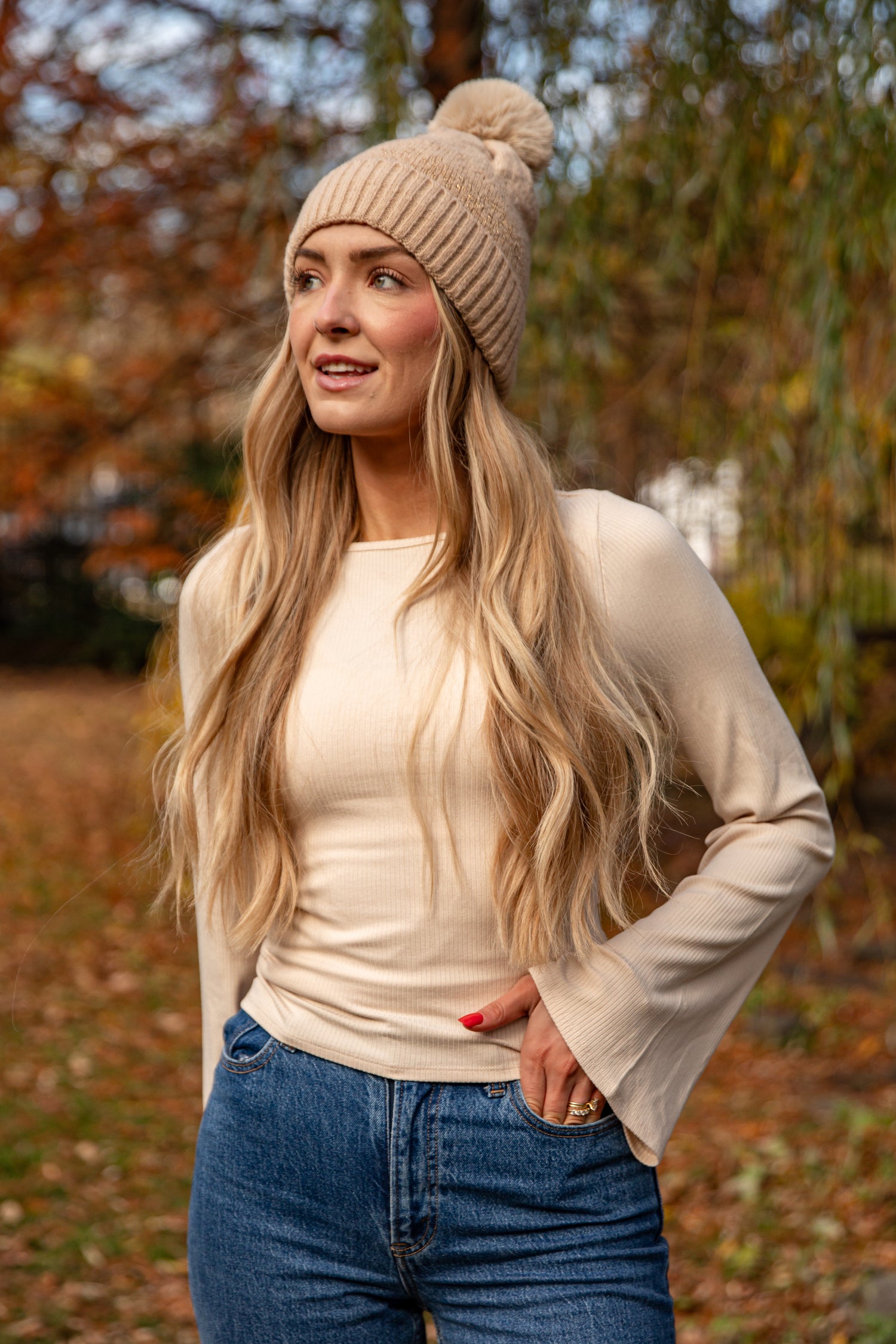 Woman wearing a beige knit beanie, cream sweater, and blue jeans standing outdoors with blurred autumn trees in the background.
