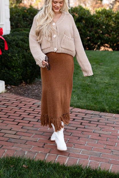Woman wearing a beige cardigan and brown skirt outdoors on a brick path.