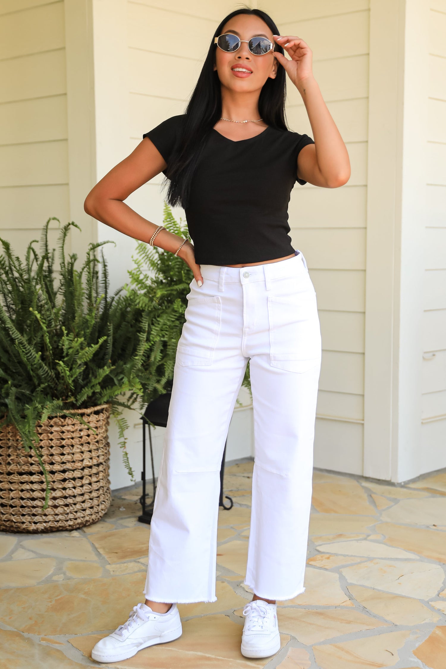 Woman wearing a black top and white pants standing on a patio with plants and a door in the background.