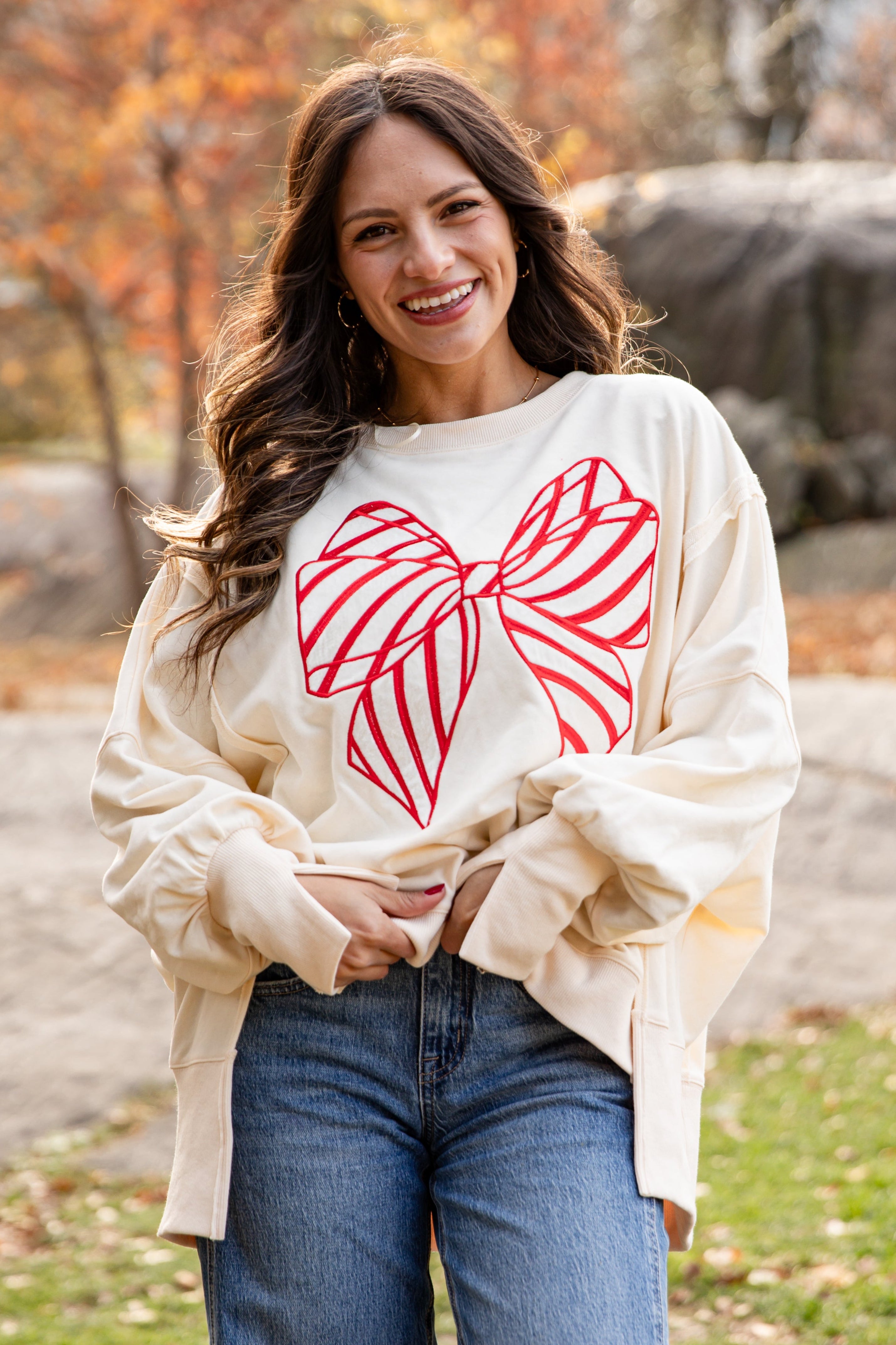 Woman wearing a cream sweatshirt with a red bow design outdoors in autumn.