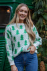 Woman wearing a sweater with green tree patterns standing next to a green vehicle decorated with Christmas trees.