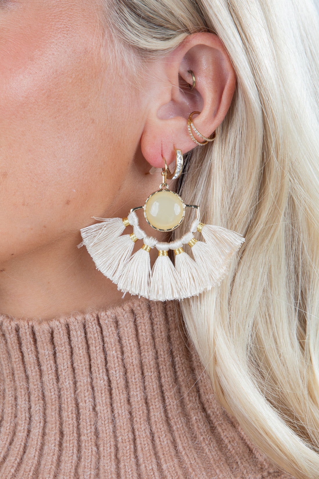 Close-up of a person wearing a gold earring with a central stone and tassel details, against a neutral background.