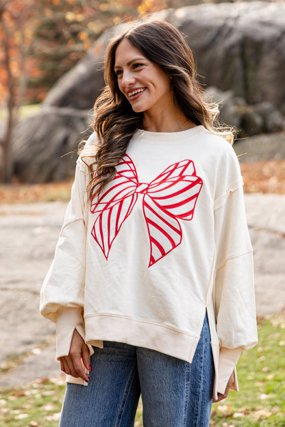 Woman wearing a cream sweater with a red bow design in an outdoor setting with trees and rocks.