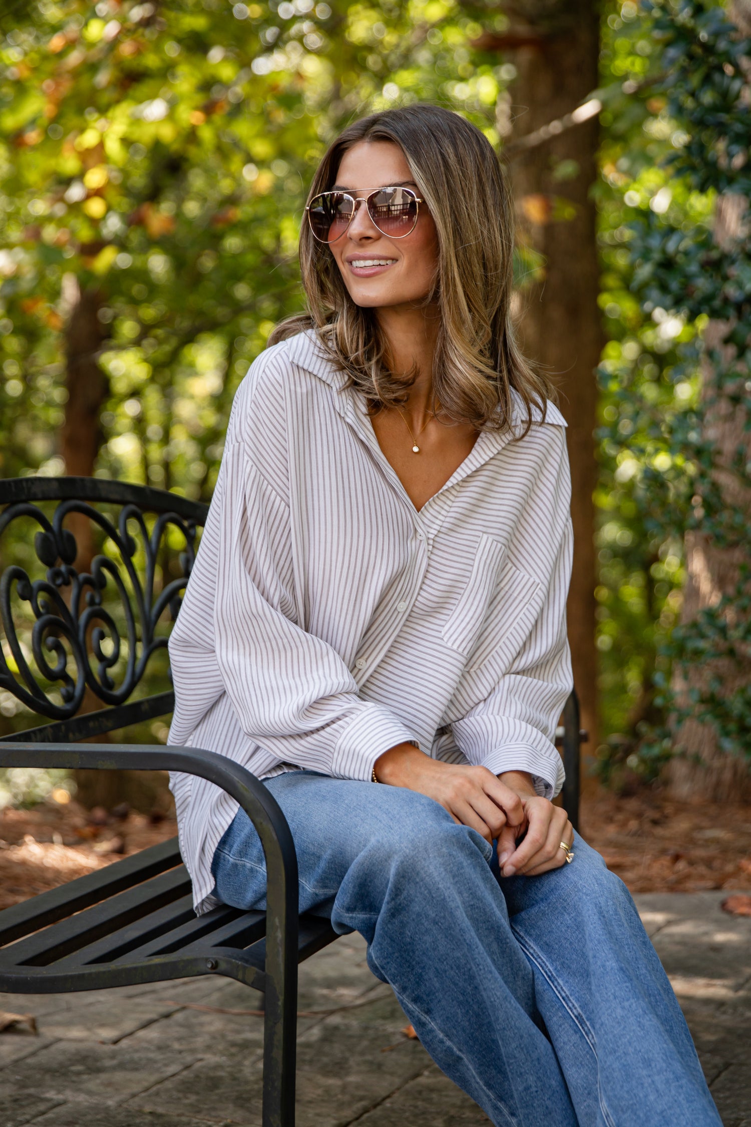 Woman sitting on a bench outdoors wearing sunglasses and a light-colored top with blue jeans.