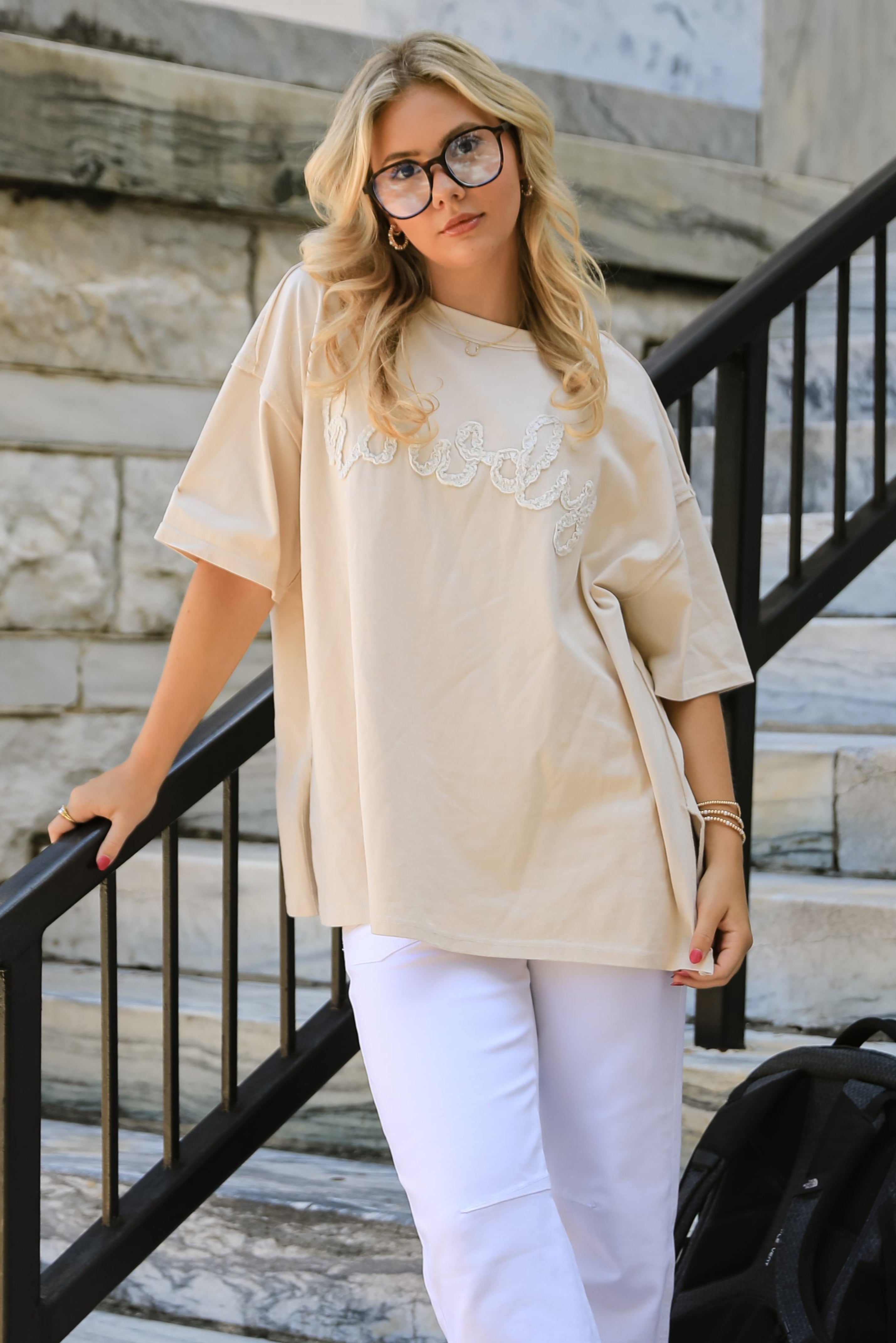 Woman standing on stone steps wearing a beige top and white pants with a black backpack.