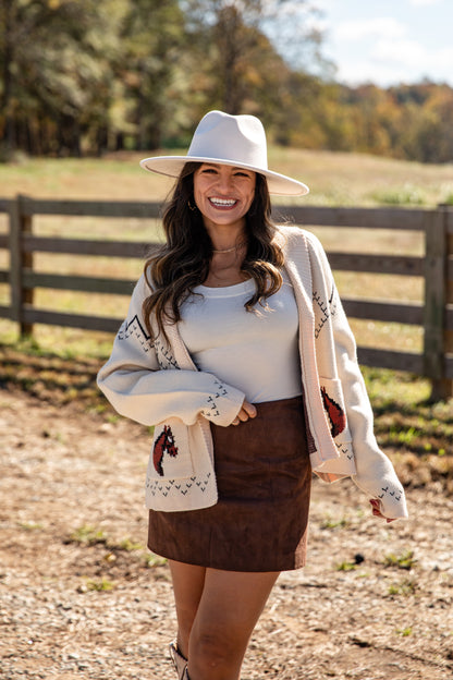 Woman wearing a white hat, white cardigan, brown skirt, and white shirt outdoors with a wooden fence and trees in the background.