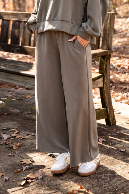 Person wearing a gray outfit with wide-leg pants sitting on a wooden bench outdoors.