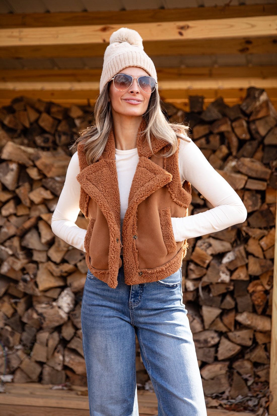 Woman wearing a brown vest, white shirt, blue jeans, and sunglasses in front of stacked firewood.