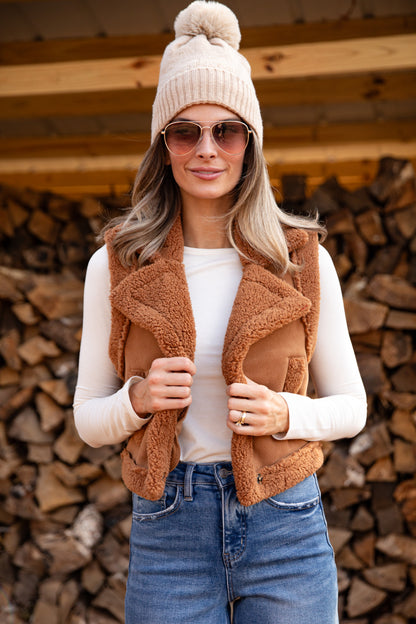 Woman wearing a beige knit hat, brown fleece vest, white shirt, and blue jeans standing in front of stacked firewood.