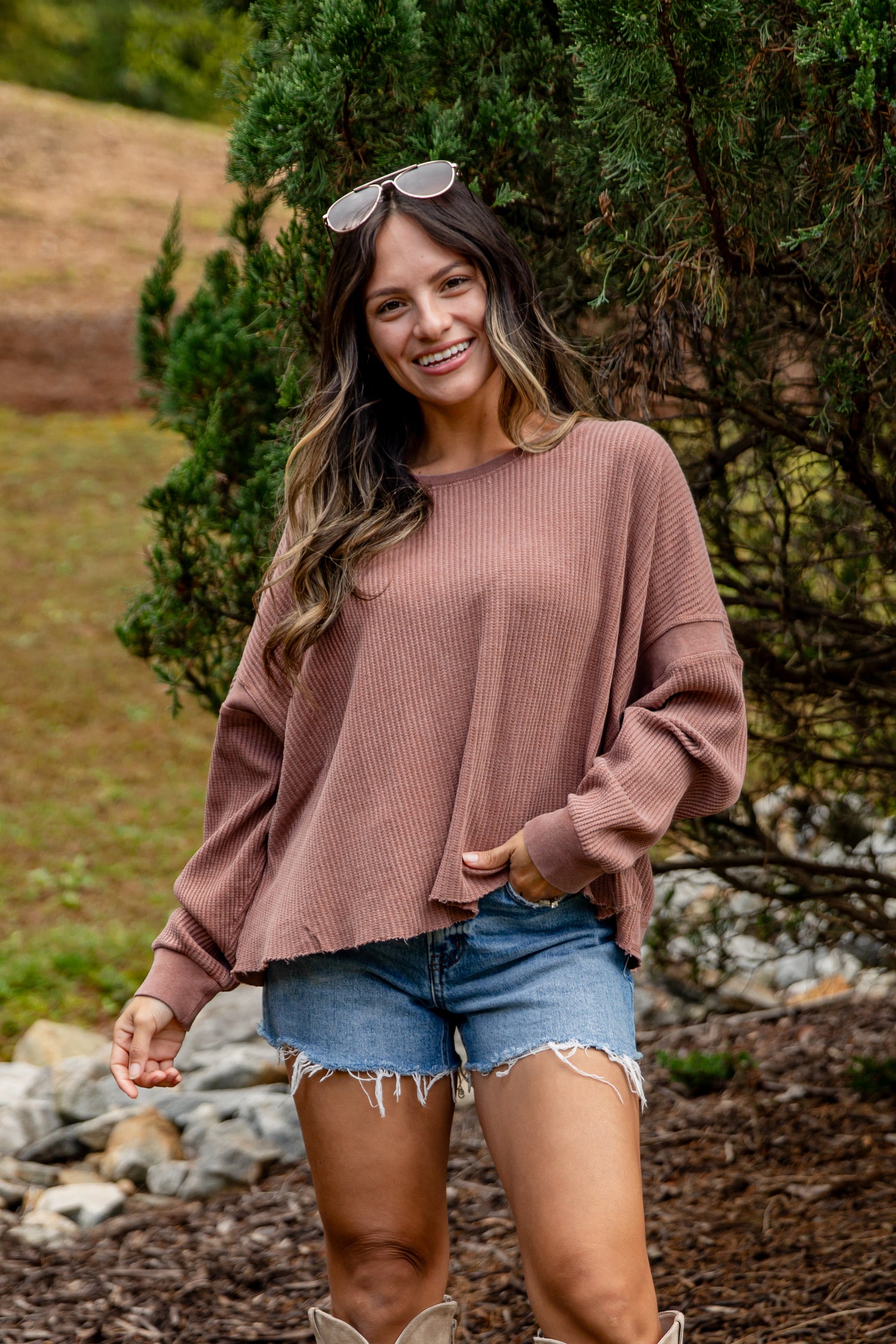 Woman wearing a pink sweater and denim shorts standing outdoors with trees and rocks in the background.