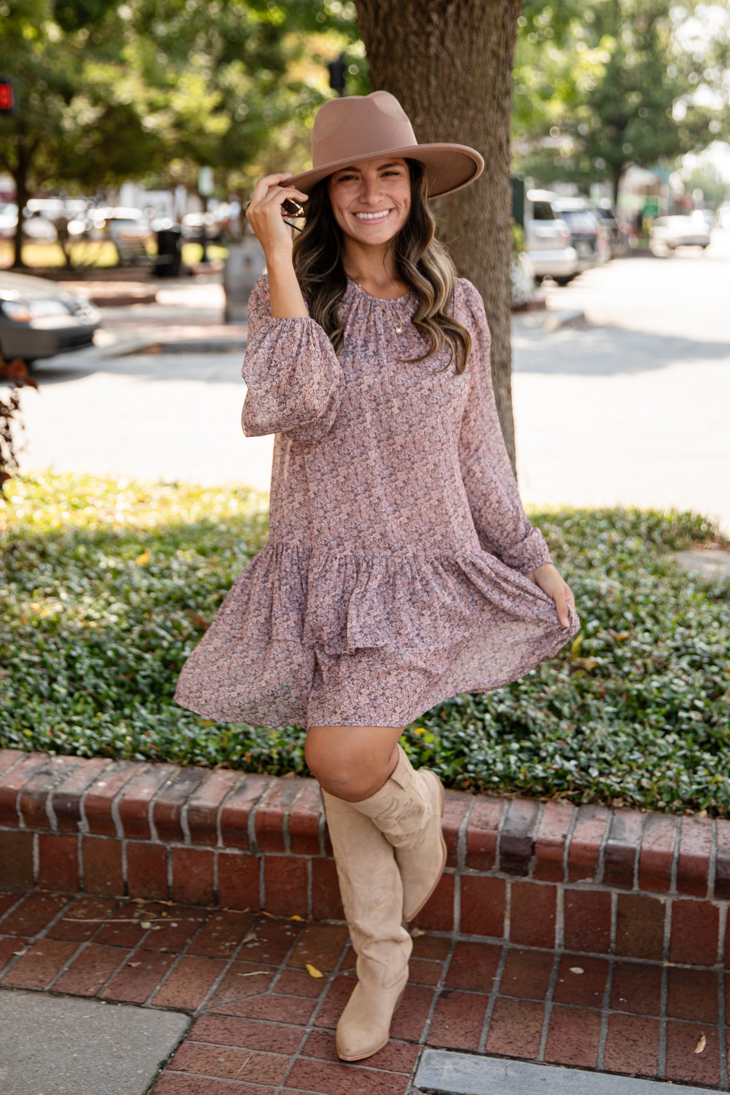 Woman in a floral dress and beige boots standing on a sidewalk with trees and cars in the background.