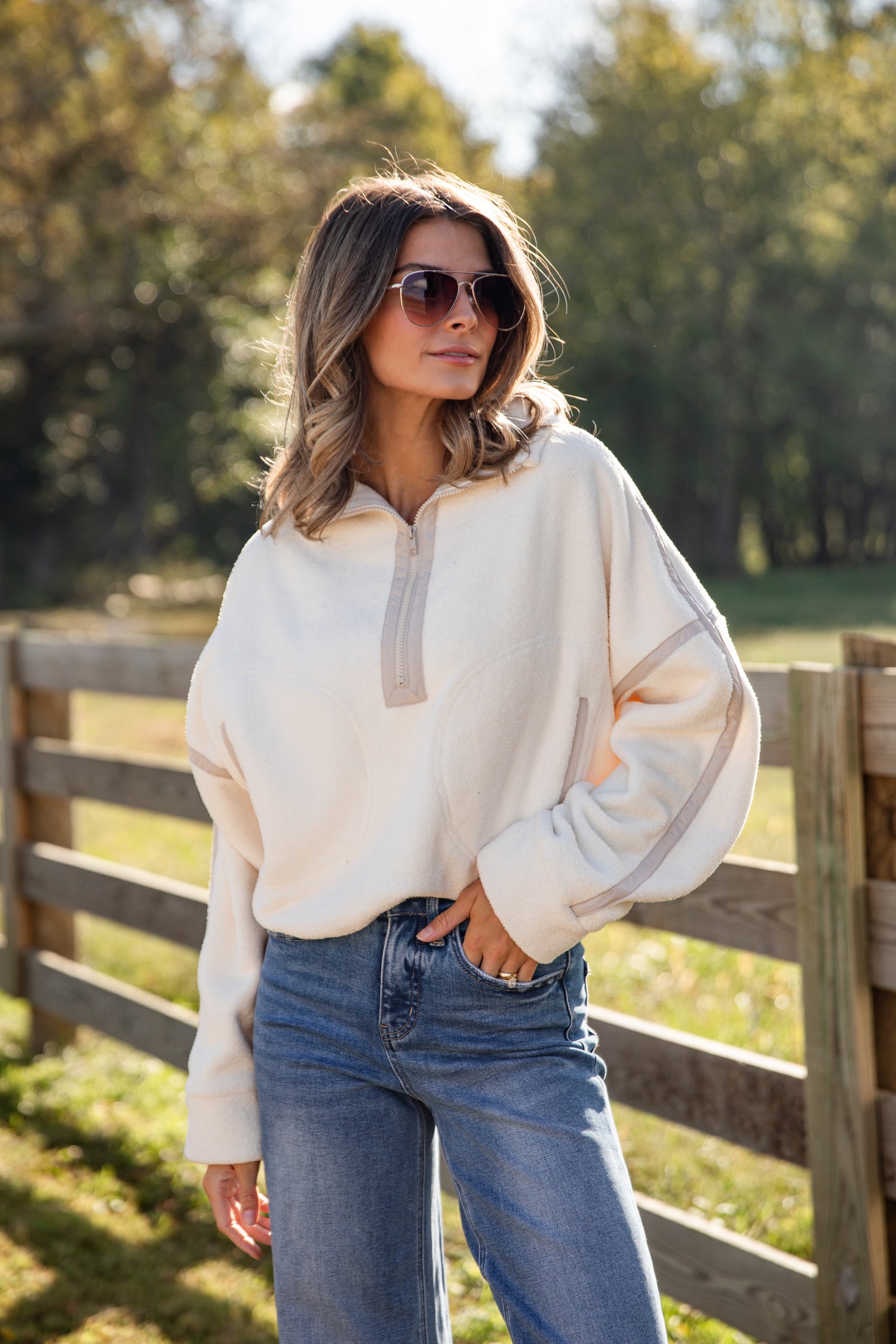 Woman wearing a cream sweater and blue jeans standing in a field with trees in the background