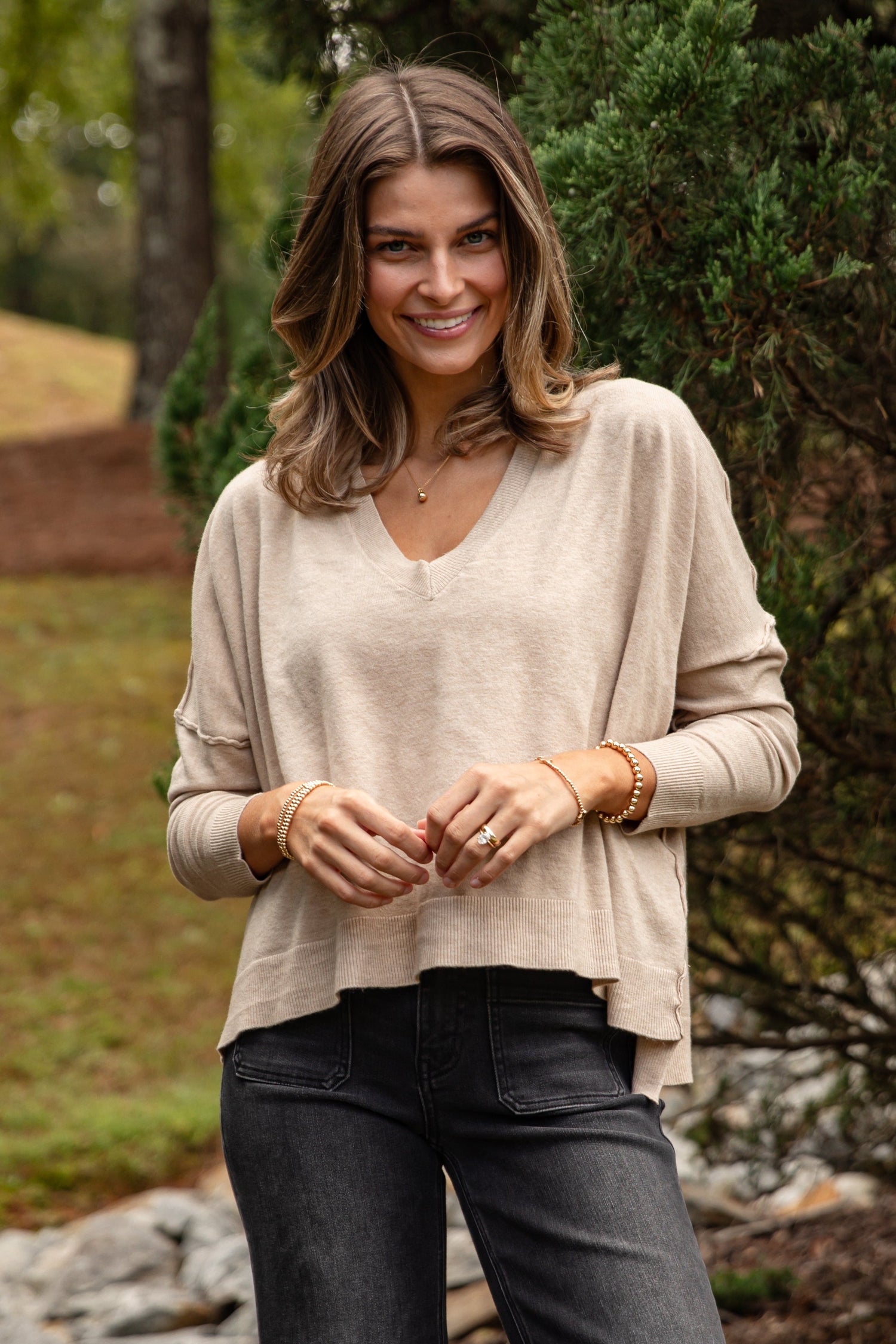 Woman wearing a beige sweater and dark jeans standing outdoors with greenery in the background