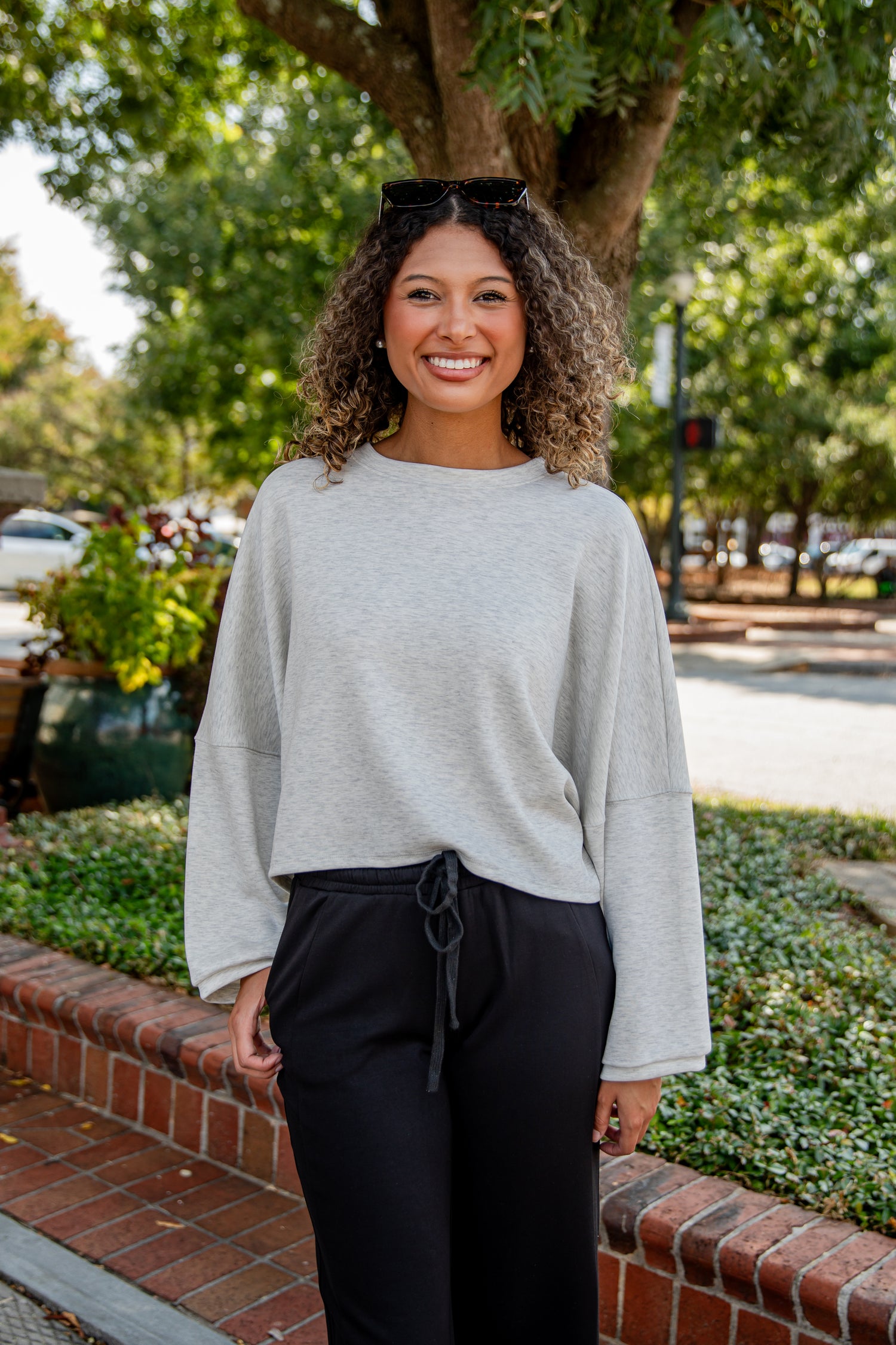Woman wearing a light gray sweater and black pants standing outdoors with trees and a street in the background.